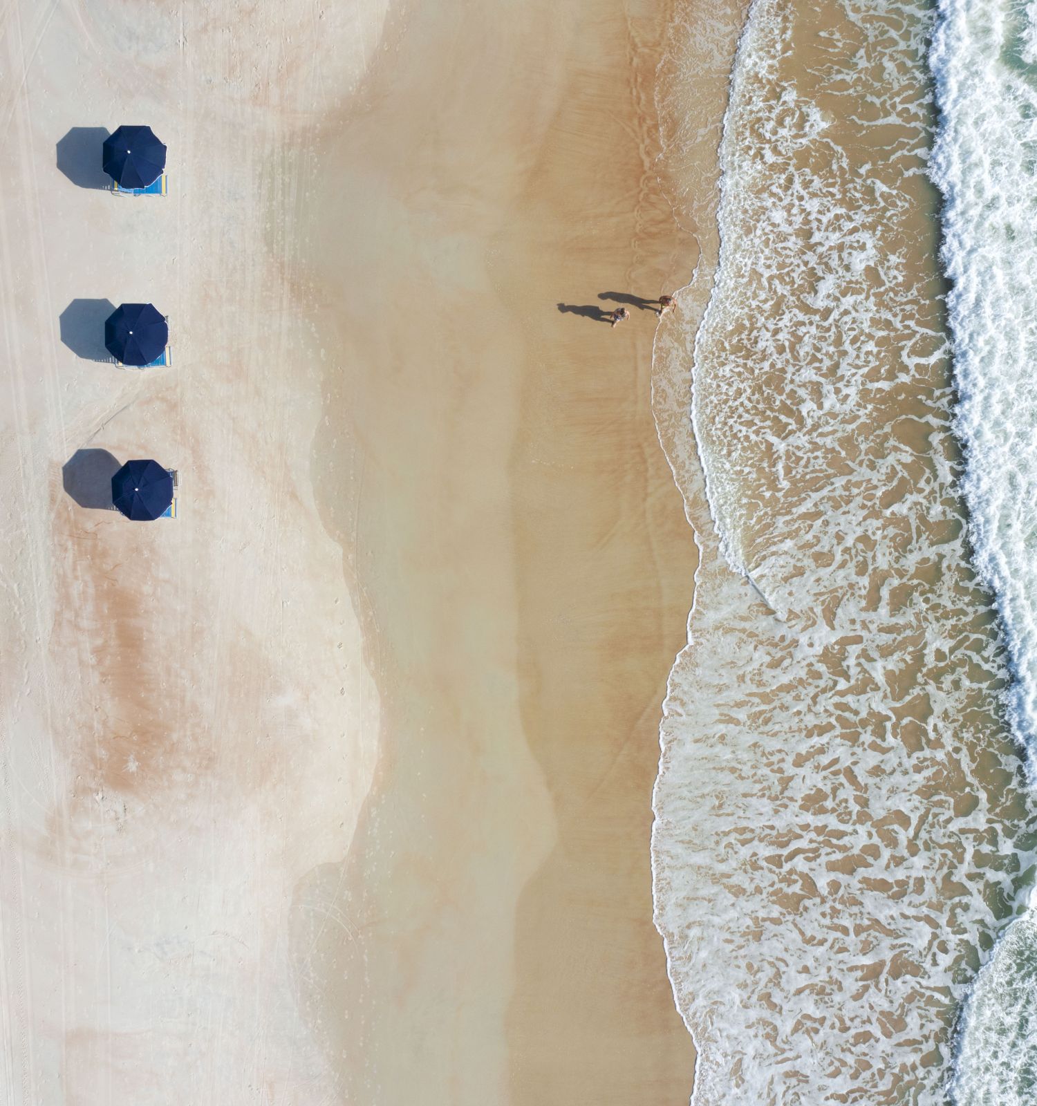 An aerial view of a beach with blue umbrellas, waves crashing on the shore, and two people walking along the wet sand.