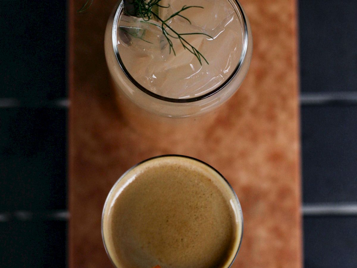 The image shows two drinks on a wooden tray: a light-colored beverage garnished with greens and a darker beverage with an orange peel garnish.