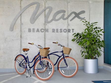 Two vintage bicycles with wicker baskets are parked near a potted plant, with the wall behind displaying the sign "Max Beach Resort."