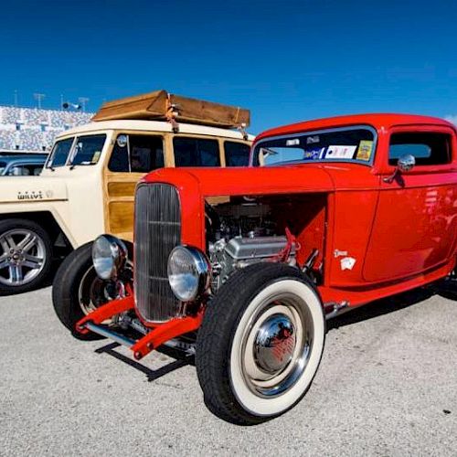 A red vintage hot rod and a classic beige car with a wooden trim are parked side by side at an outdoor event under a clear blue sky.