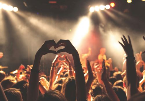 A crowd at a concert with hands raised and forming heart shapes, illuminated by stage lights and performing musicians in the background.