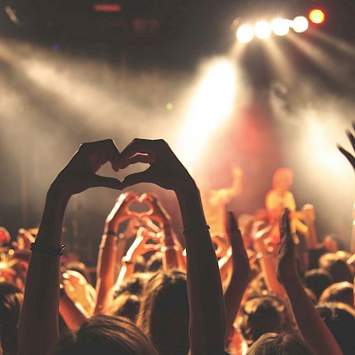 A crowd at a concert with hands raised and forming heart shapes, illuminated by stage lights and performing musicians in the background.