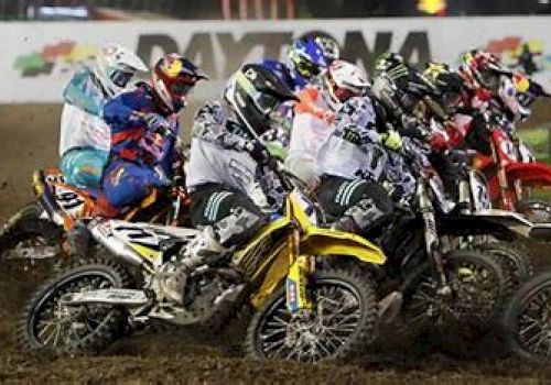 The image shows a group of motocross riders competing on a dirt track, with the word 