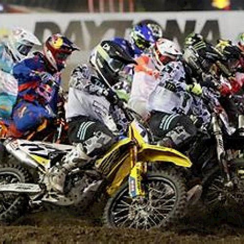 The image shows a group of motocross riders competing on a dirt track, with the word 