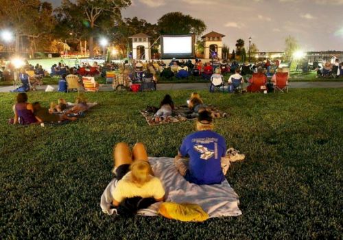 People sitting on a lawn watching an outdoor movie at night. Lights illuminate the area and trees can be seen in the background.