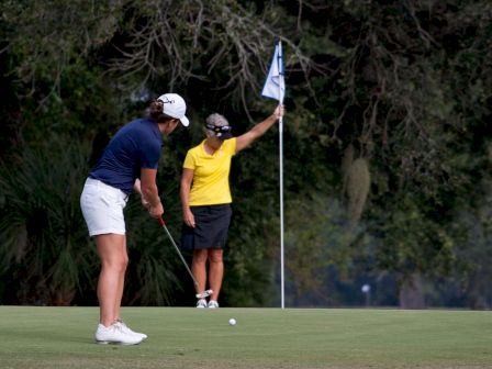 Two people are on a golf green, one putting while the other holds the flagstick near the hole, amidst a backdrop of trees.