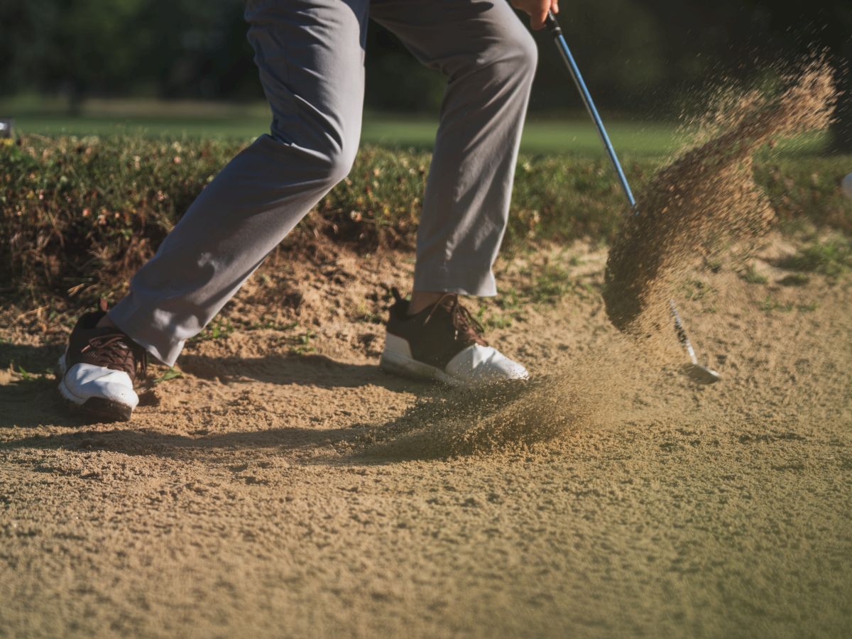 A golfer is hitting a golf ball out of a sand bunker, with sand flying up as the club makes contact. Trees are blurry in the background.