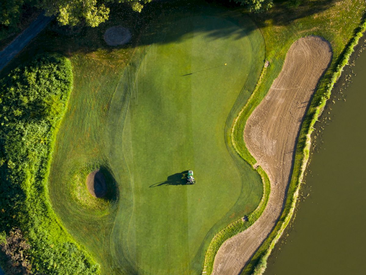 An aerial view of a golf green featuring a sand trap, surrounding trees, and adjacent water hazard.