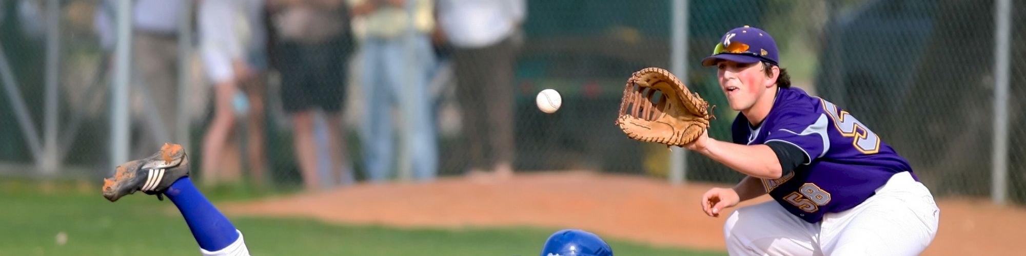 A baseball player in a blue uniform slides into a base as another player in a purple uniform prepares to catch the ball with an outstretched glove.