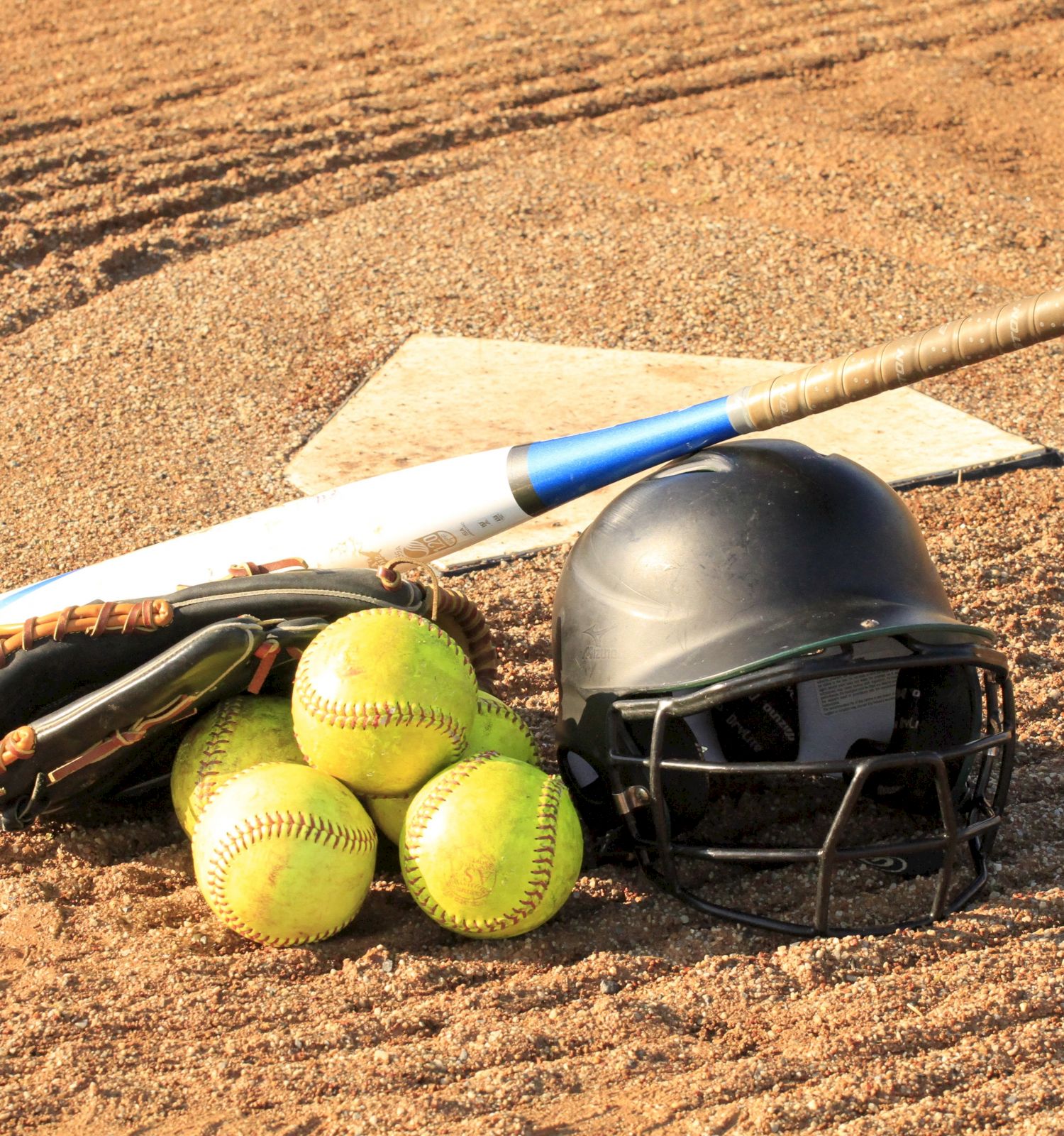 This image shows a baseball bat, a glove, four softballs, and a helmet on a dirt field with a home plate in the background.