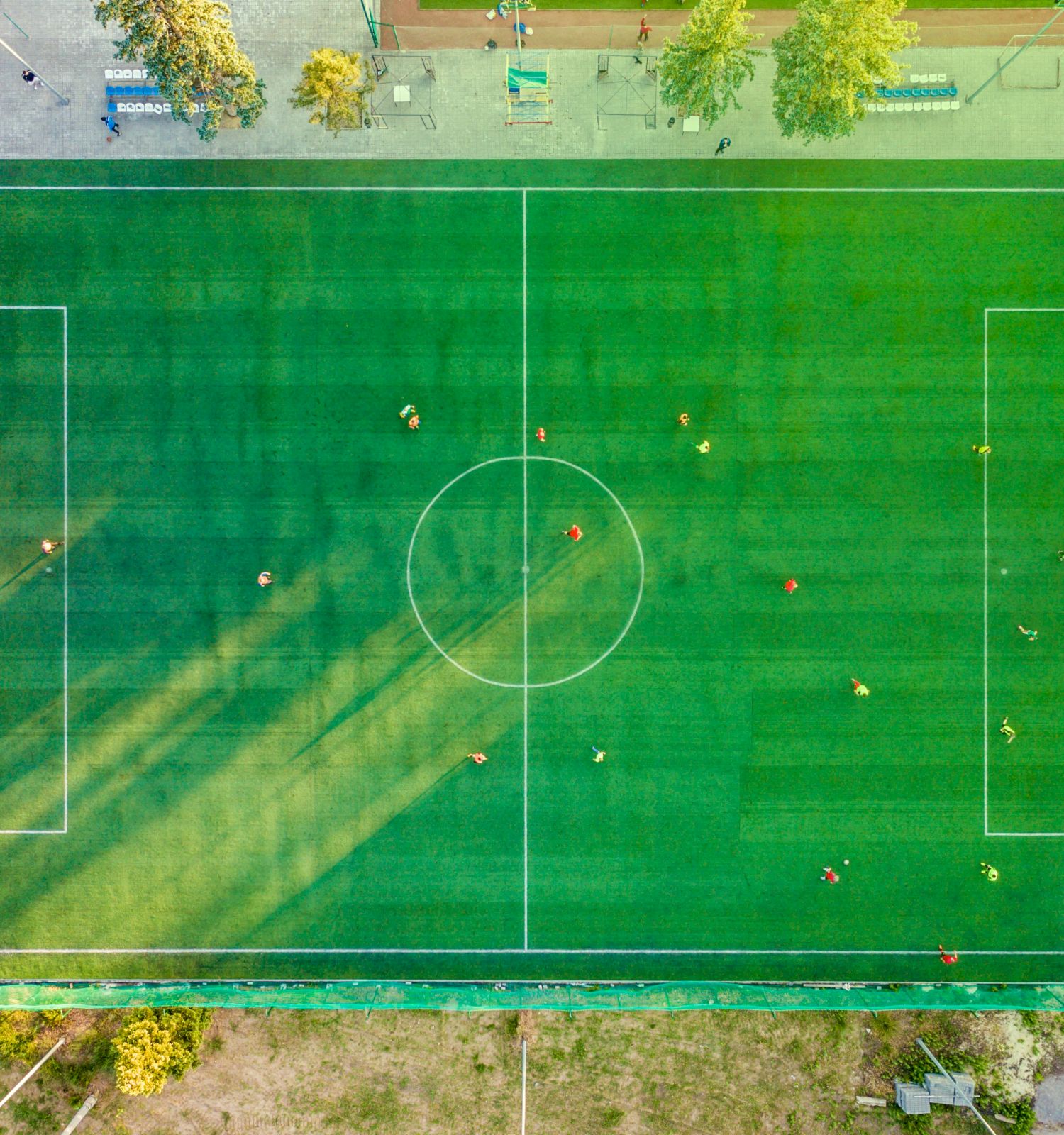 An aerial view of a football field with players on it, surrounded by greenery and adjacent structures, under the sunlight.