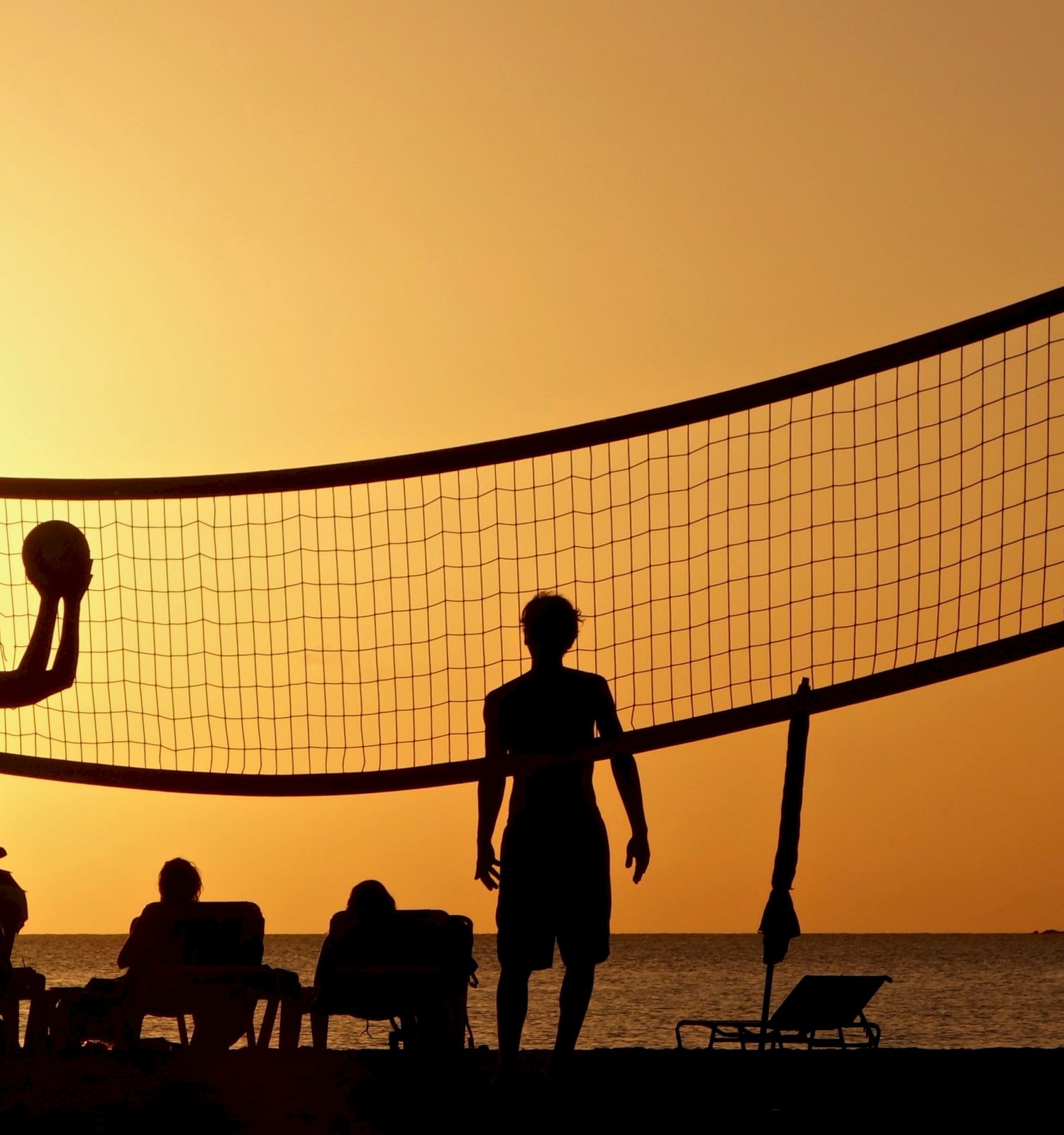 Silhouettes of people playing volleyball on a beach during sunset, with a net and relaxed onlookers in the background.