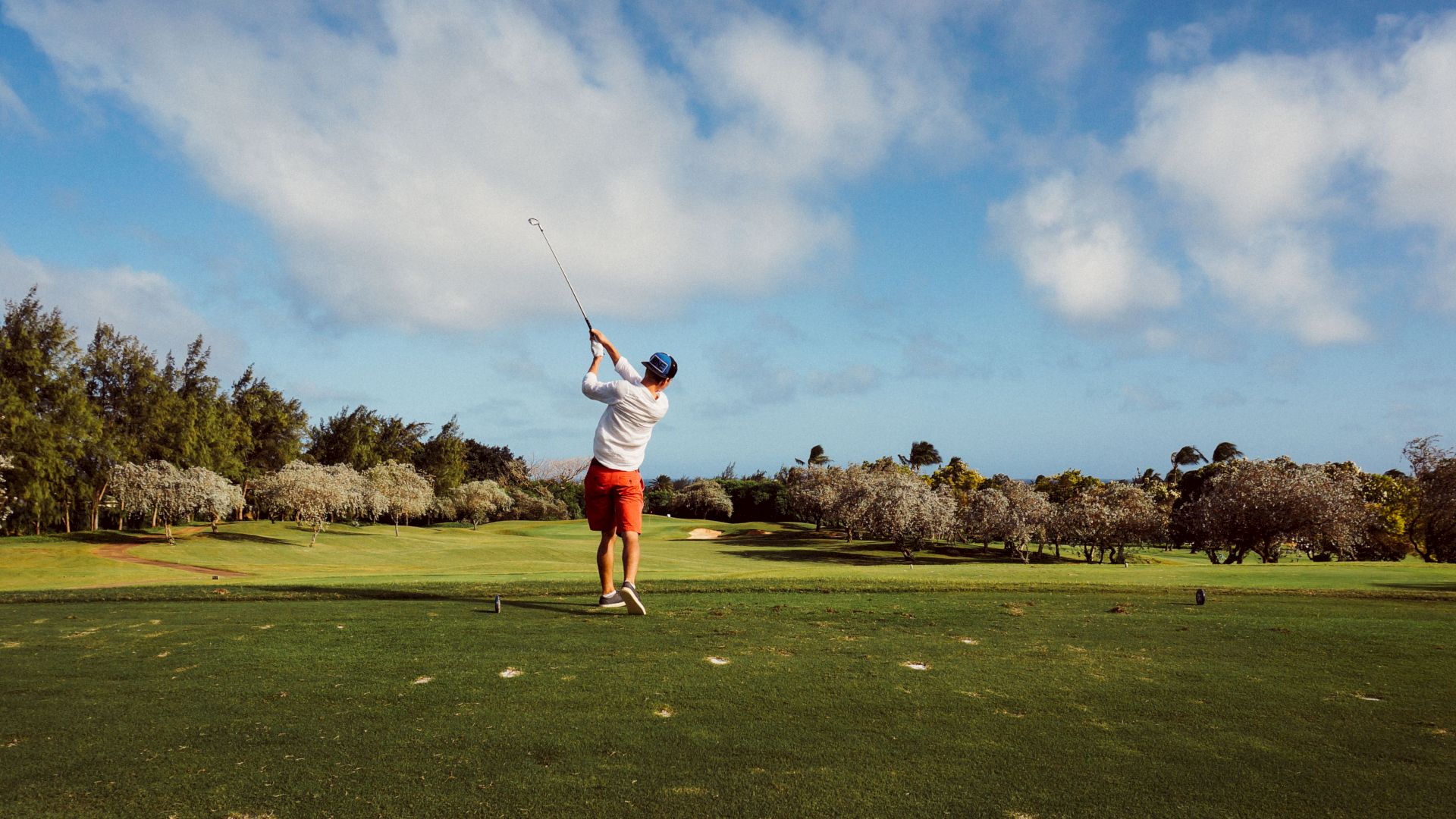 A person is mid-swing on a golf course with lush greenery and trees in the background under a partly cloudy sky.