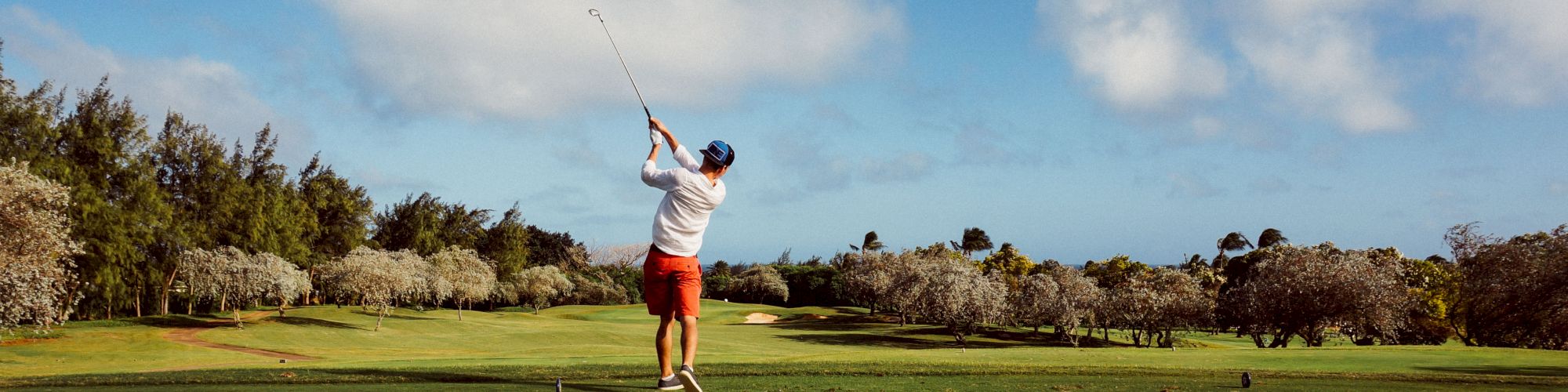 A person is mid-swing on a golf course with lush greenery and trees in the background under a partly cloudy sky.