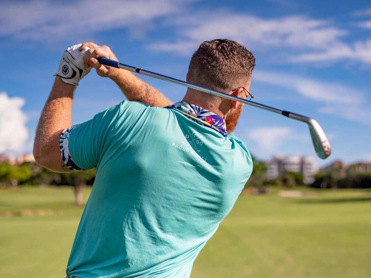 A person in a turquoise shirt is swinging a golf club on a golf course, with a scenic background of grass, trees, and a partly cloudy sky.