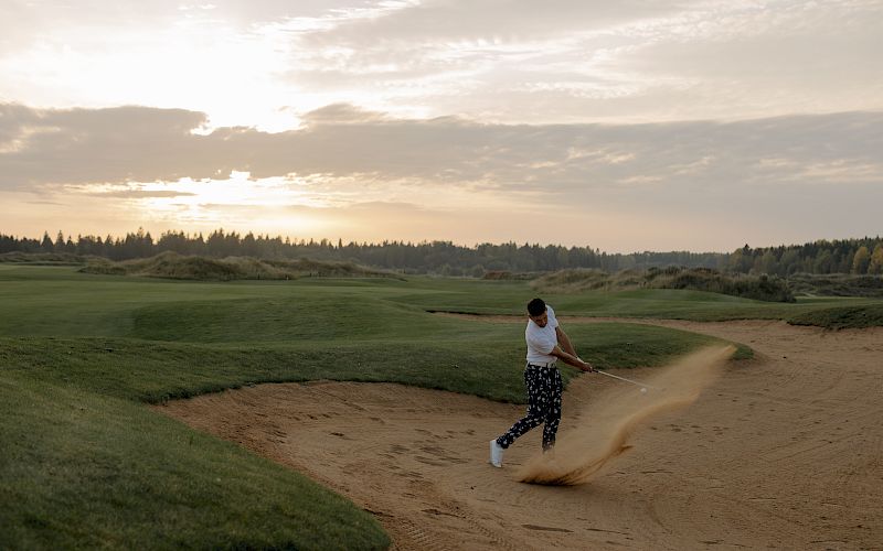 A golfer is hitting a shot from a sand bunker on a golf course during sunset, with sand flying around.