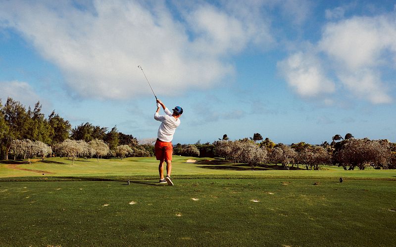 A person is playing golf on a lush green course, swinging a club under a partly cloudy sky, surrounded by trees and open landscape.