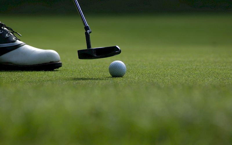 A golf ball is positioned in front of a putter on a green, with a golfer's shoe visible in the background. The scene is set on a golf course.