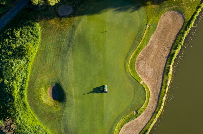 An aerial view of a golf course green surrounded by sand bunkers and adjacent to a water hazard, with trees bordering the area.