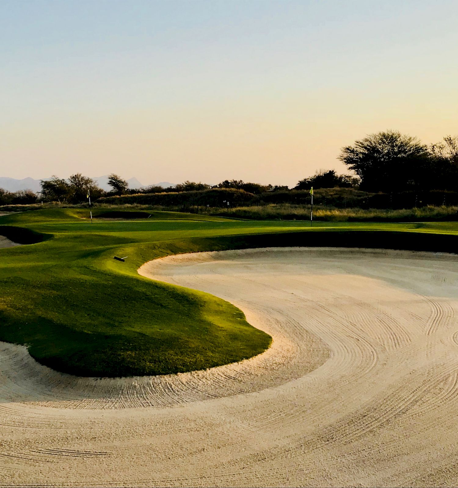 A golf course with green fairways and multiple sand bunkers under a clear sky, surrounded by distant trees and small hills in the background.