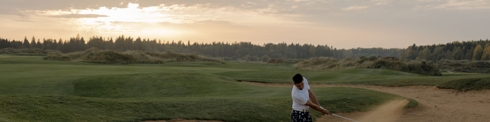 A person is playing golf, hitting a ball out of a sand trap on a lush green golf course under a partly cloudy sky at sunset.