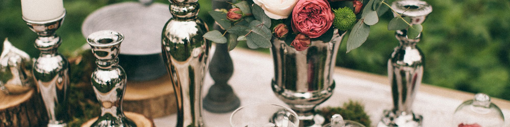 The image shows a decorated table with candles, silver candlesticks, a floral arrangement with roses, glassware, and rustic wooden accents.