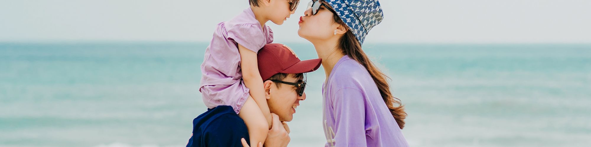 A family is enjoying a day at the beach. The father is carrying a child on his shoulders, and the mother is giving the child a kiss. The sea is in the background.