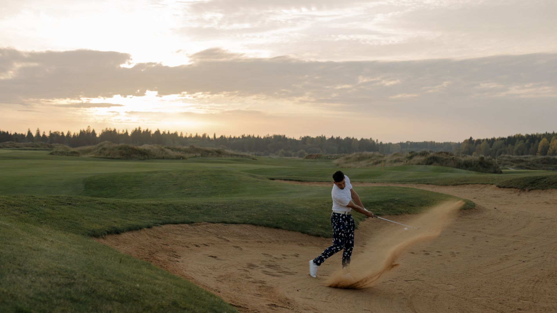 A golfer in patterned pants hits a shot with sand flying from a bunker on a golf course during sunset, with a scenic background of trees and sky.