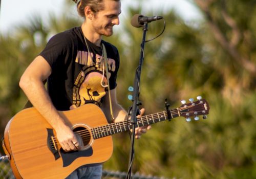 A person is playing an acoustic guitar outdoors, smiling, with a microphone stand nearby, wearing a black T-shirt and jeans by a fenced area with greenery.