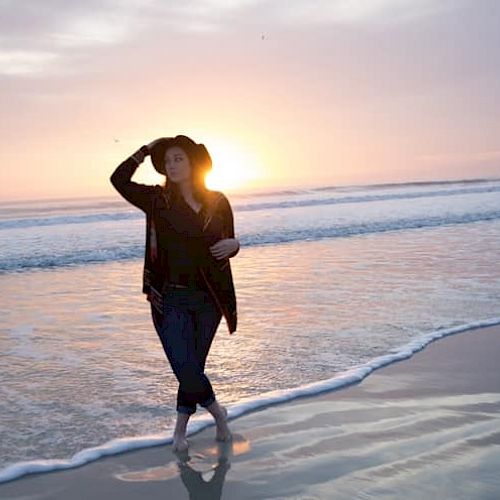 A person in casual attire walks along a beach at sunset, with gentle waves rolling in, creating a serene and peaceful atmosphere.