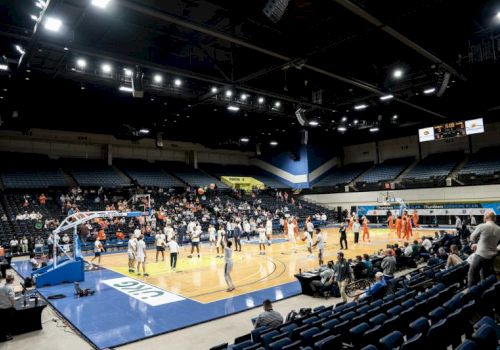 The image shows a basketball game taking place in an indoor arena with players and spectators.