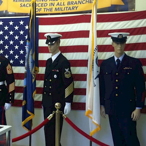 Three uniformed military personnel stand in front of large American flags, roped off in a display. They wear dress uniforms with medals and caps.