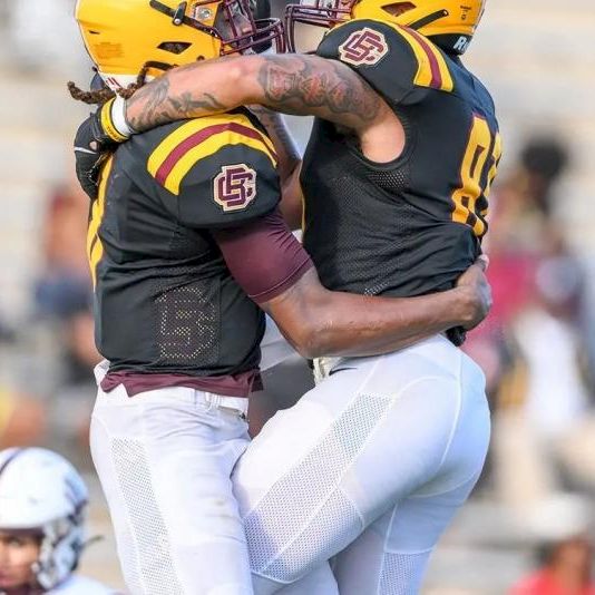 Two football players in black uniforms hug mid-field, celebrating a score while other players and blurred crowd watch in the background.