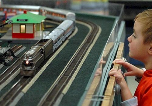 A child watches a model train set with tracks, a train, and a small building inside a glass display.