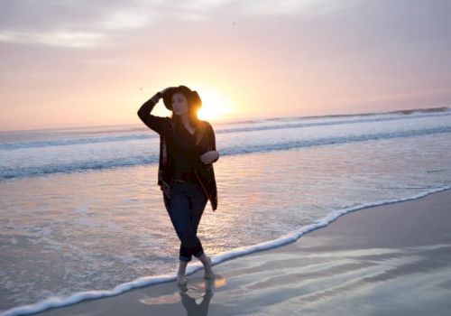 A person walking along a beach at sunset, wearing a hat and light clothing, with the waves gently lapping at the shore's edge.