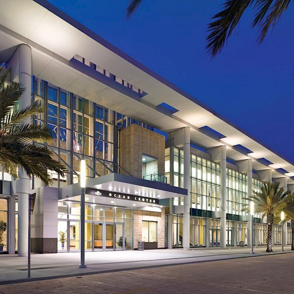 A modern hotel corridor at dusk/night with palm trees lining the walkway, illuminated windows, and an &ldquo;OCEAN CLUB&rdquo; entrance.