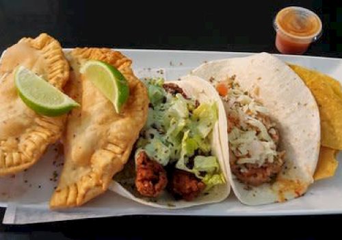 The image shows empanadas with lime garnish, two tacos with various fillings, tortilla chips, and a small cup of sauce, served on a plate.