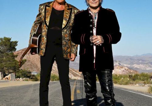 Two men stand on a desert road with mountains behind them&mdash;one in a gold patterned jacket carrying a guitar, the other in black holding hands.