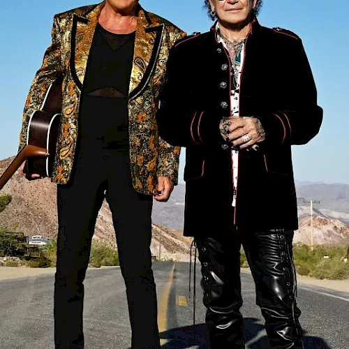 Two men stand on a desert road with mountains behind them&mdash;one in a gold patterned jacket carrying a guitar, the other in black holding hands.
