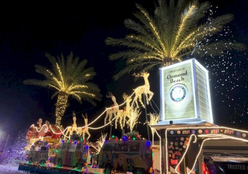 A brightly lit parade float at night with palm trees and a sign reading &ldquo;Welcome to Ormond Beach&rdquo;.