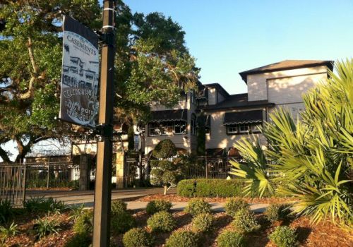The image shows a charming building surrounded by trees and shrubs with a sign in the foreground, under a clear blue sky.