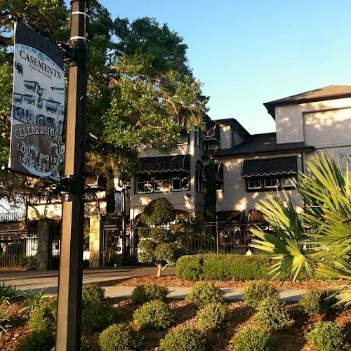 The image shows a charming building surrounded by trees and shrubs with a sign in the foreground, under a clear blue sky.