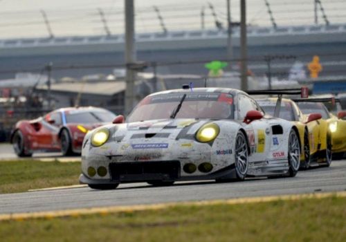 A white-and-red race car with headlights on leads other GT cars on a track near the pit lane.