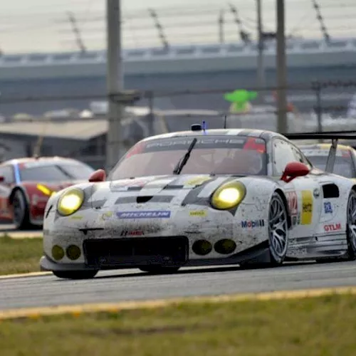 A white-and-red race car with headlights on leads other GT cars on a track near the pit lane.