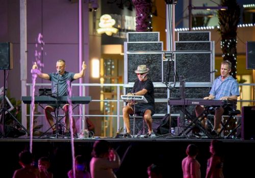 Three musicians perform on stage with keyboards, colorful lighting, and an audience below, creating a lively atmosphere.