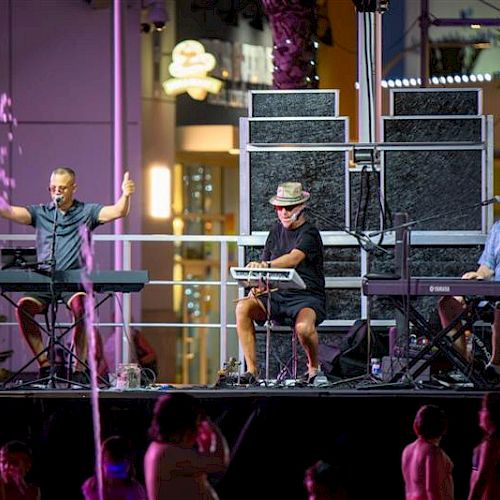 Three musicians perform on stage with keyboards, colorful lighting, and an audience below, creating a lively atmosphere.