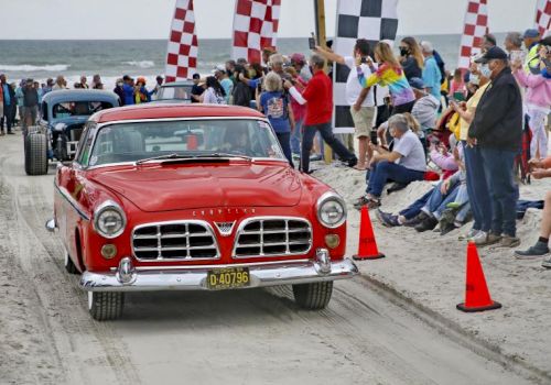 A red vintage Chevrolet leads a beach parade as crowds cheer and wave checkered flags near the ocean.