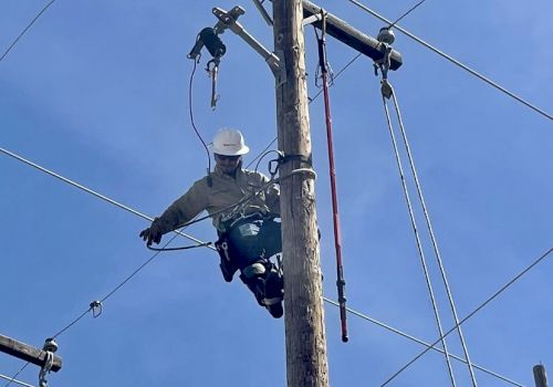 A person wearing safety gear is climbing a utility pole, seemingly performing maintenance or repairs on the electrical wires with a clear blue sky background.