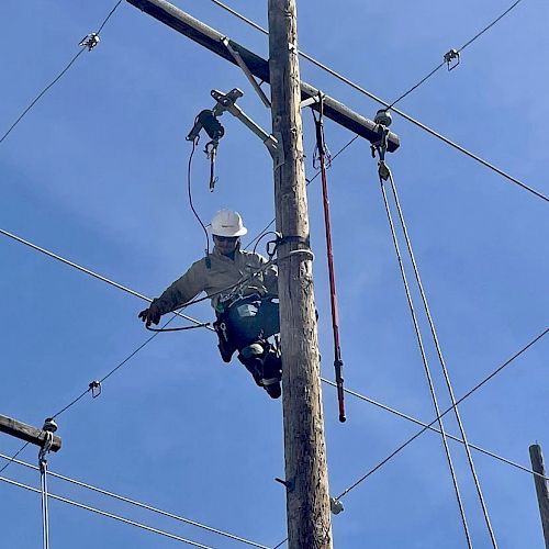 A person wearing safety gear is climbing a utility pole, seemingly performing maintenance or repairs on the electrical wires with a clear blue sky background.