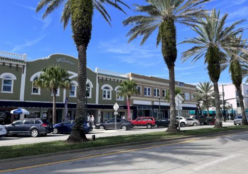 A street scene with palm trees, parked cars, people walking, and a row of colorful buildings under a clear blue sky.